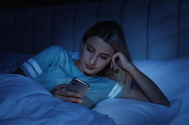 Девушка лежит в постели глядя в экран смартфона в темноте