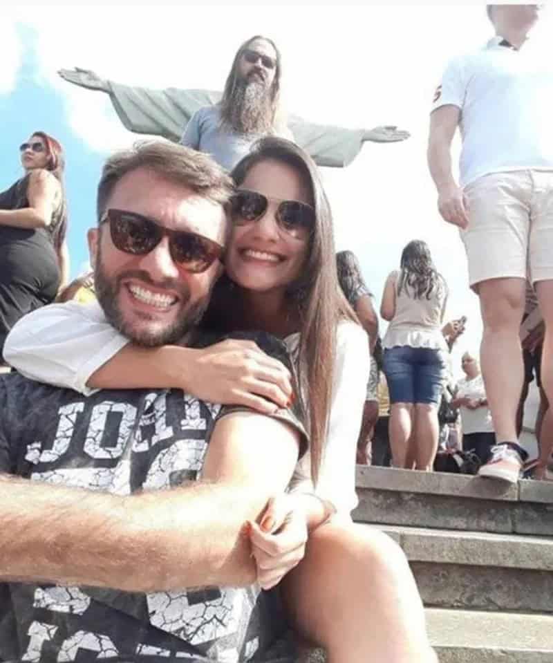 Пара обнимается у статуи Христа в Рио-де-Жанейро, мужчина за ними случайно повторяет жест раскинутых рук