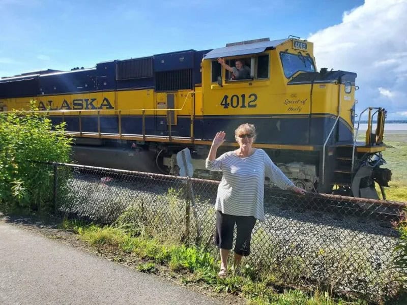 Женщина машет рукой на фоне локомотива Alaska Railroad, машинист выглядывает из окна кабины