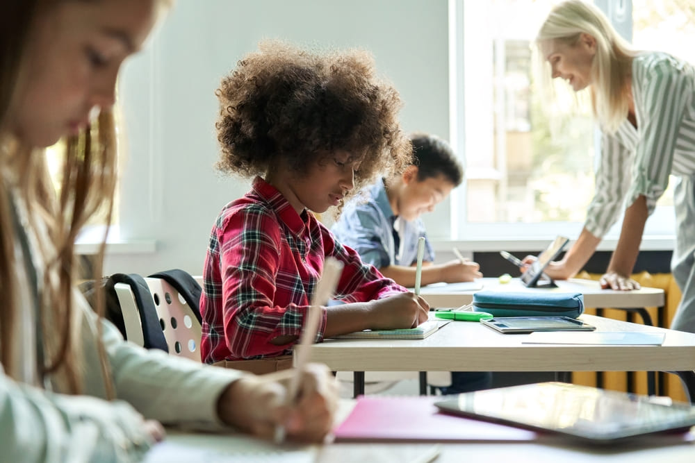 Diligent concentrated hard working diverse miltiethnic classmates schoolchildren writing tasks in notebooks with happy teacher explaining and helping. Schoolwork in diversity team. Education concept.