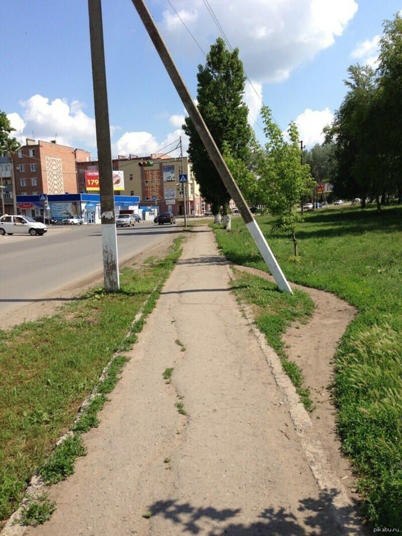 Тротуар с сильно наклонённым столбом и деревьями на фоне